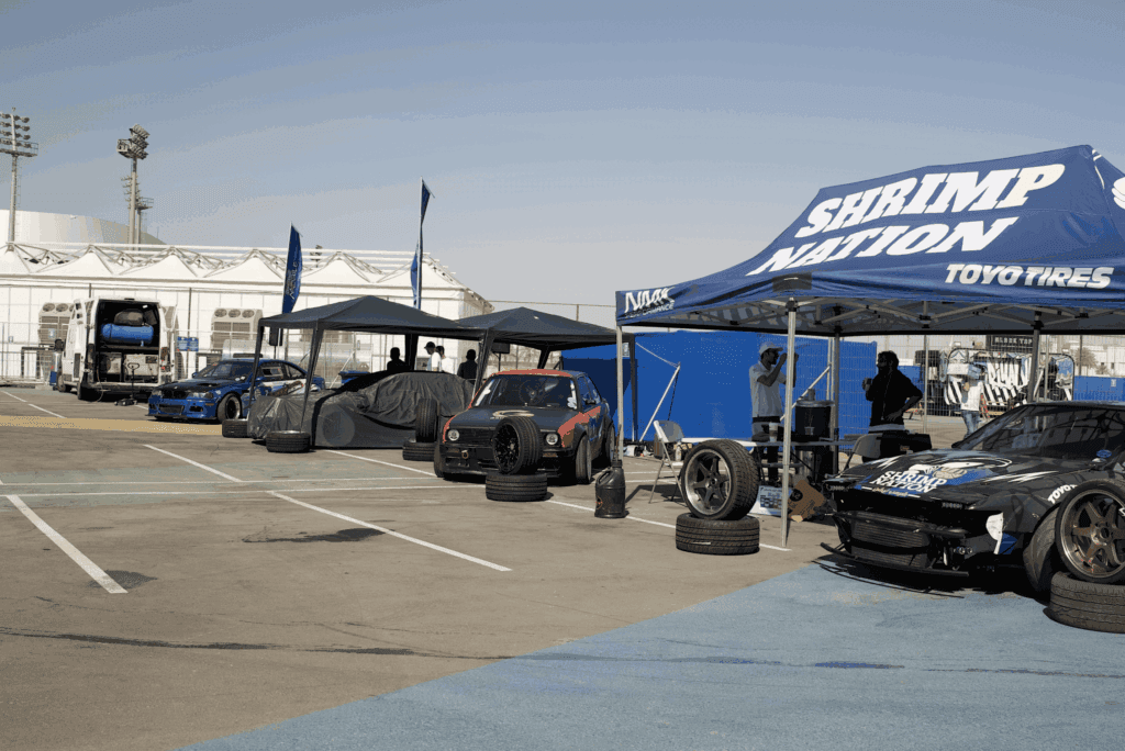 A pit area featuring drift cars and a tent labeled 'Shrimp Nation' at a motorsport event, showcasing image.