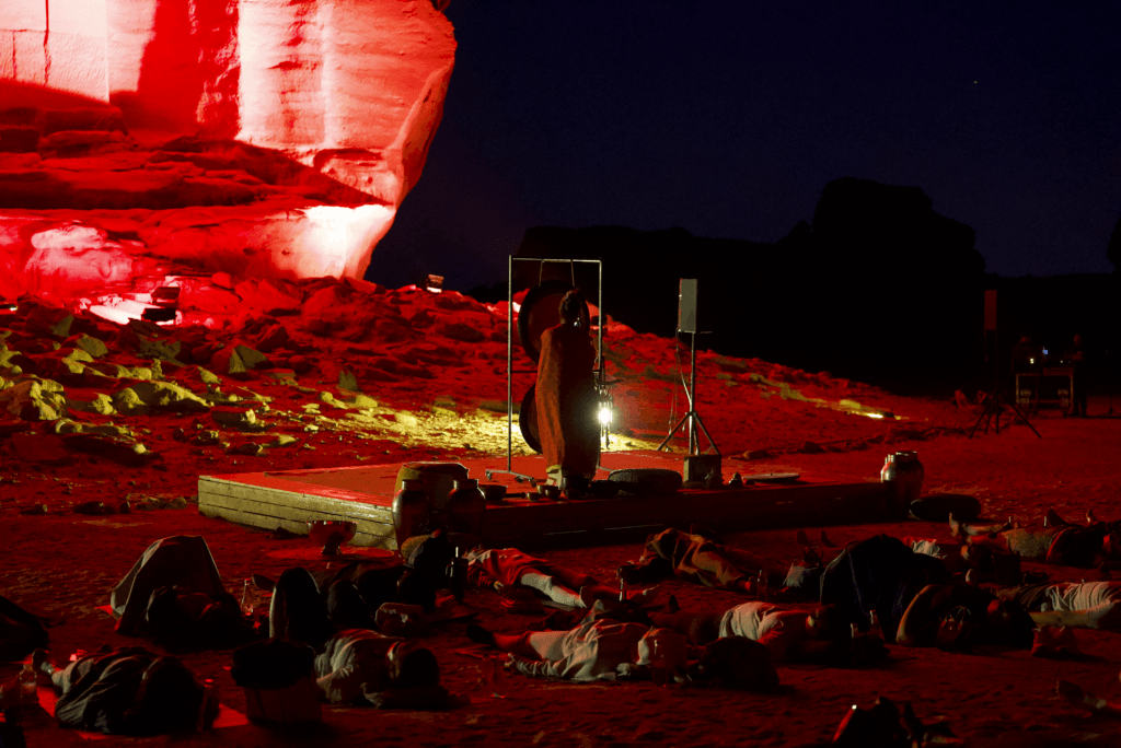 A red-lit performance in the desert at night, with people lying on the ground and a performer on stage, showcasing image.
