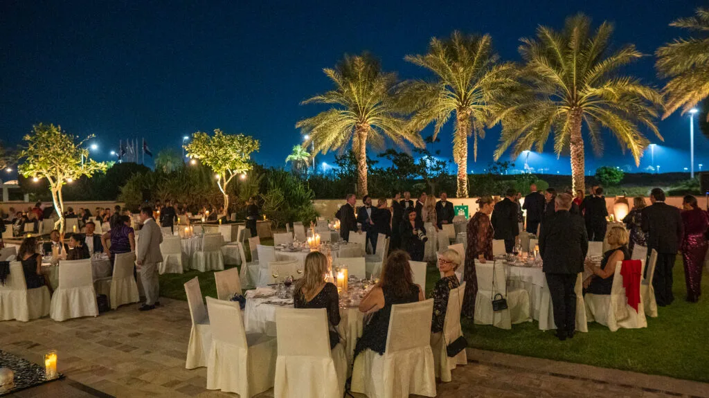 An outdoor evening event with dining tables surrounded by palm trees, showcasing image.