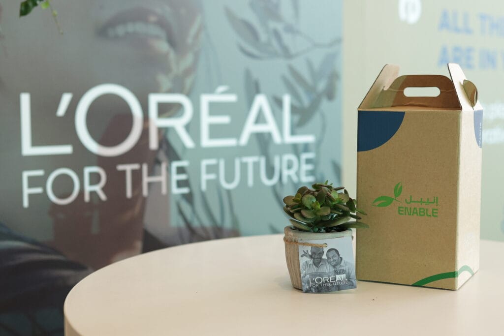 A small plant in a wooden pot next to a cardboard box, with a L'Oreal for the Future backdrop, showcasing image.