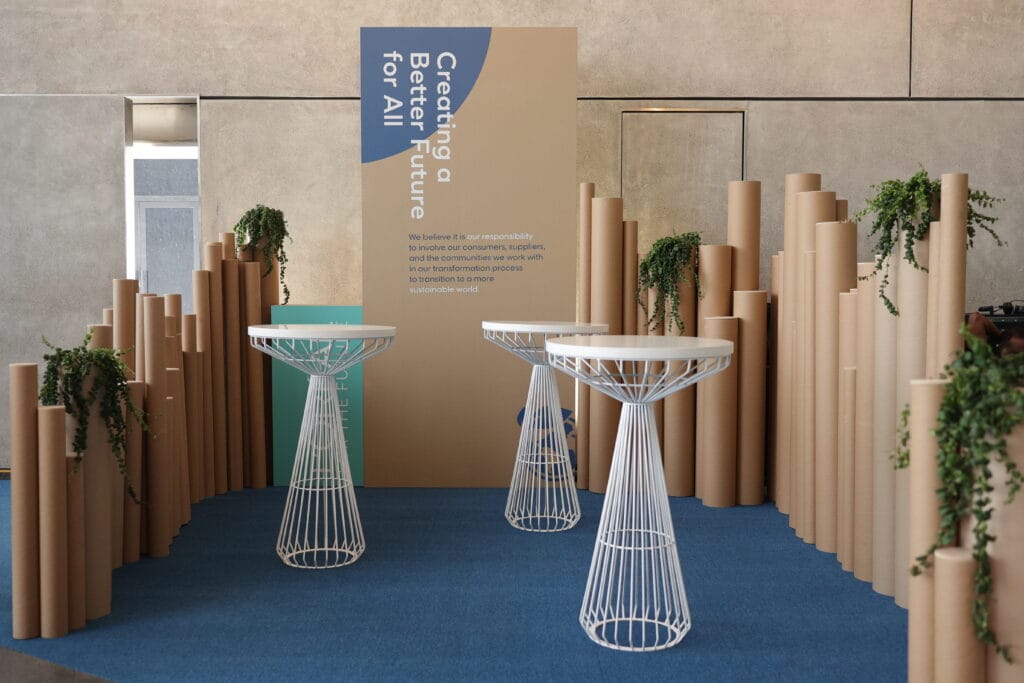 A modern event space featuring white tables surrounded by cardboard columns and greenery, showcasing image.