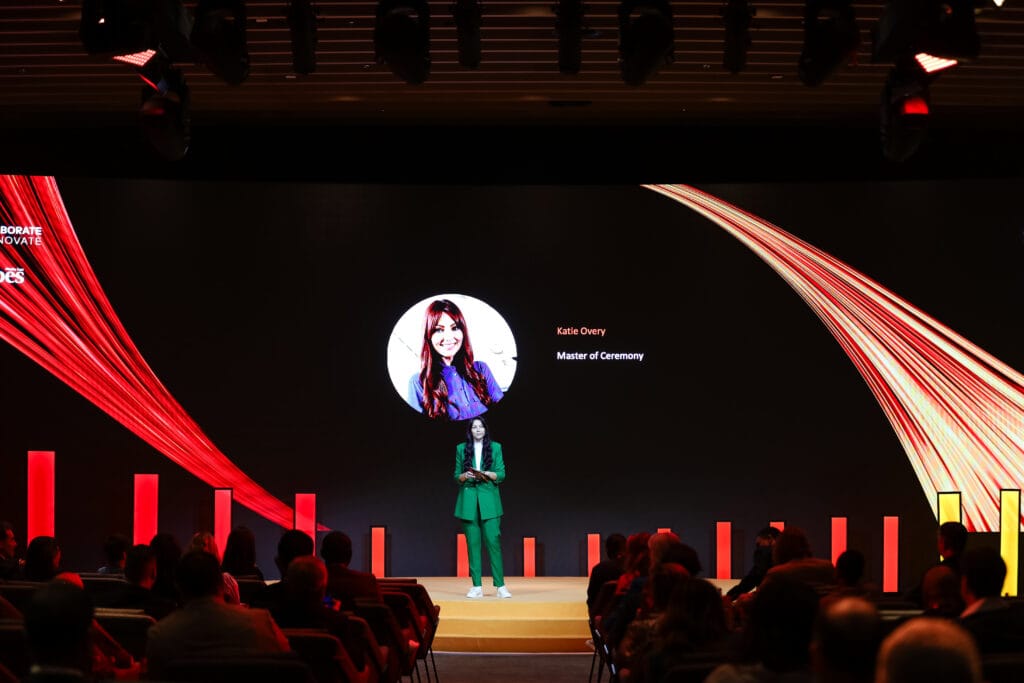 Katie Overy as Master of Ceremony presenting on stage with a colorful backdrop, showcasing image.