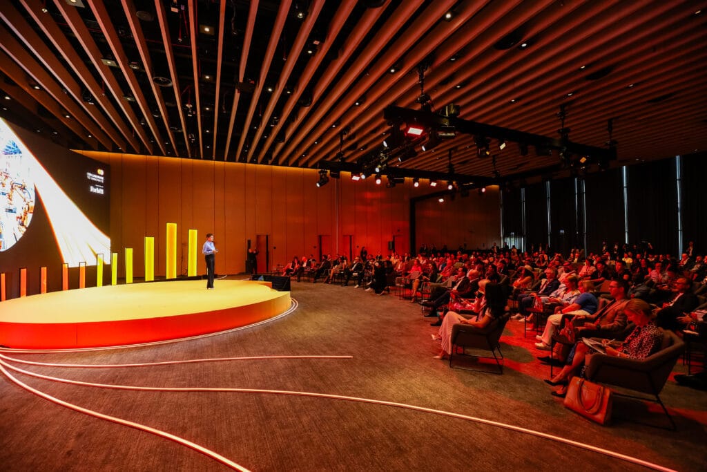 A speaker presenting on stage in a conference hall with a large audience, showcasing image.