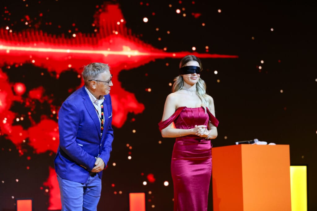 A blindfolded woman in a red dress stands on stage during a performance, highlighting image.