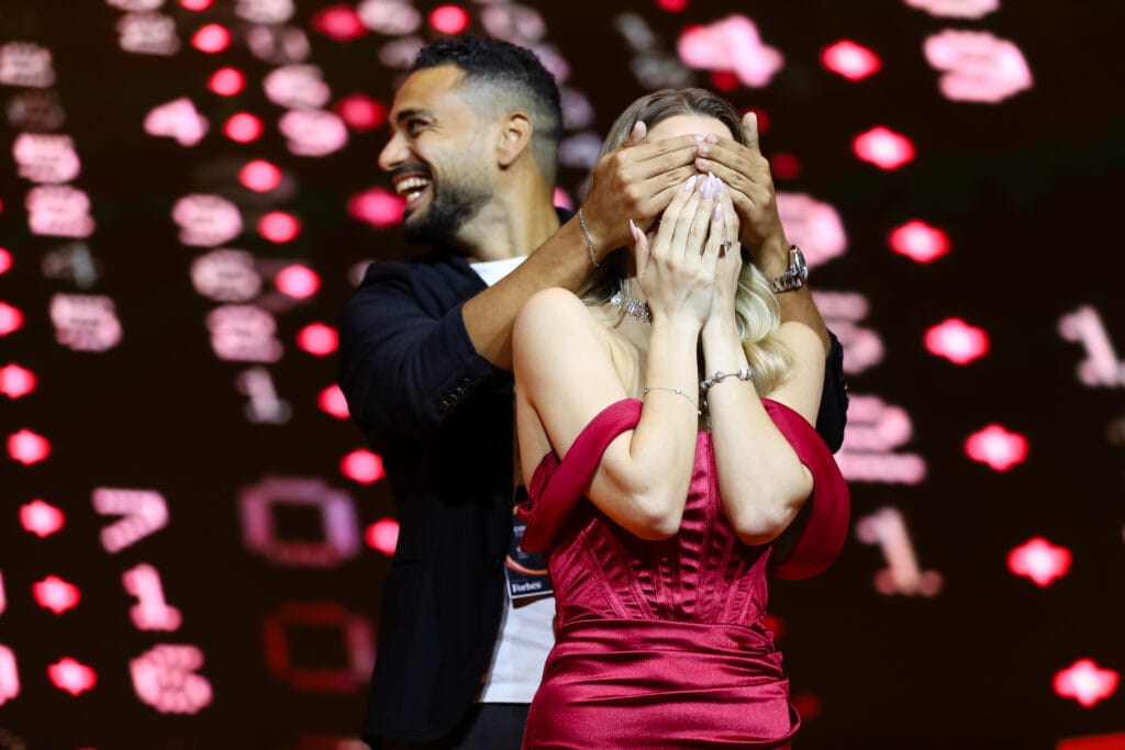 A man playfully covering a woman's eyes on stage, she is wearing a red dress, highlighting image.