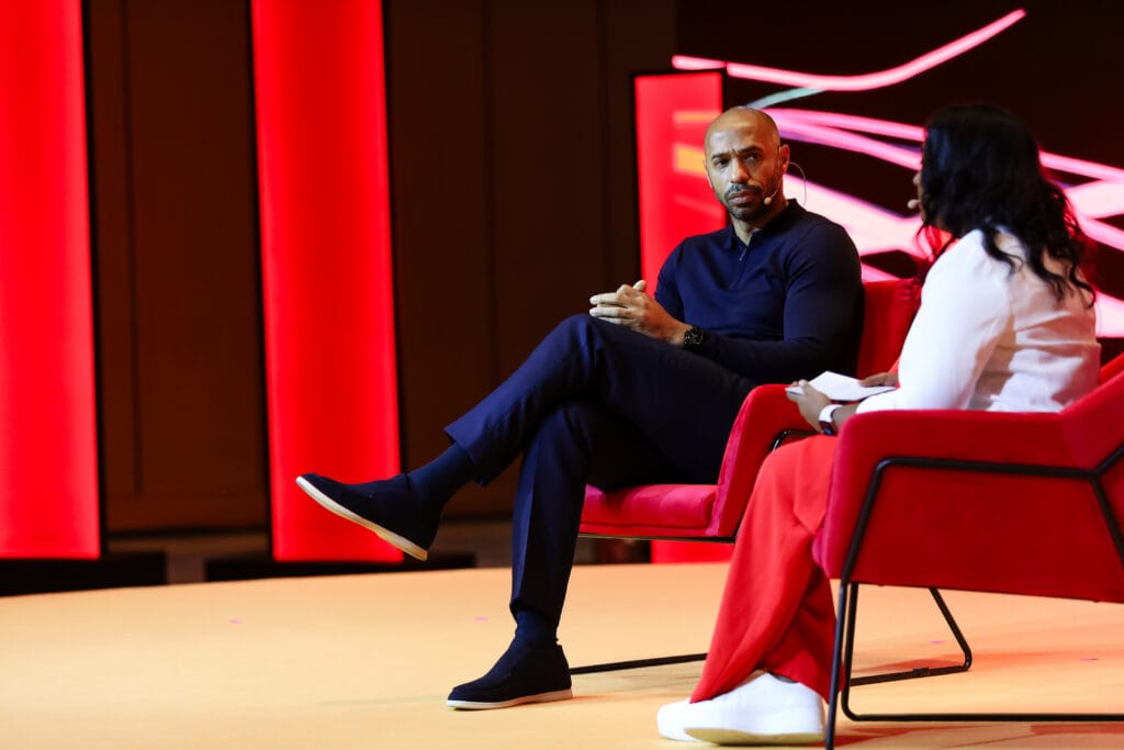 A man and woman seated on red chairs engaged in a discussion on stage, highlighting image.