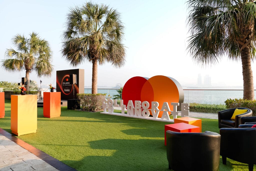 An outdoor event setup featuring palm trees, colorful podiums, and a sign that reads 'Collaborate to Innovate', showcasing image.